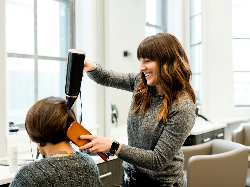 A hairdresser laughing with a client while blow-drying her hair in a bright salon
